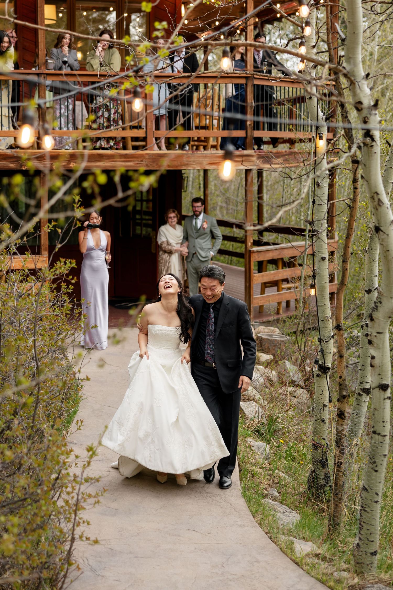 Joyful bride in white gown laughs with groom on garden path at rustic woodland wedding venue with string lights.