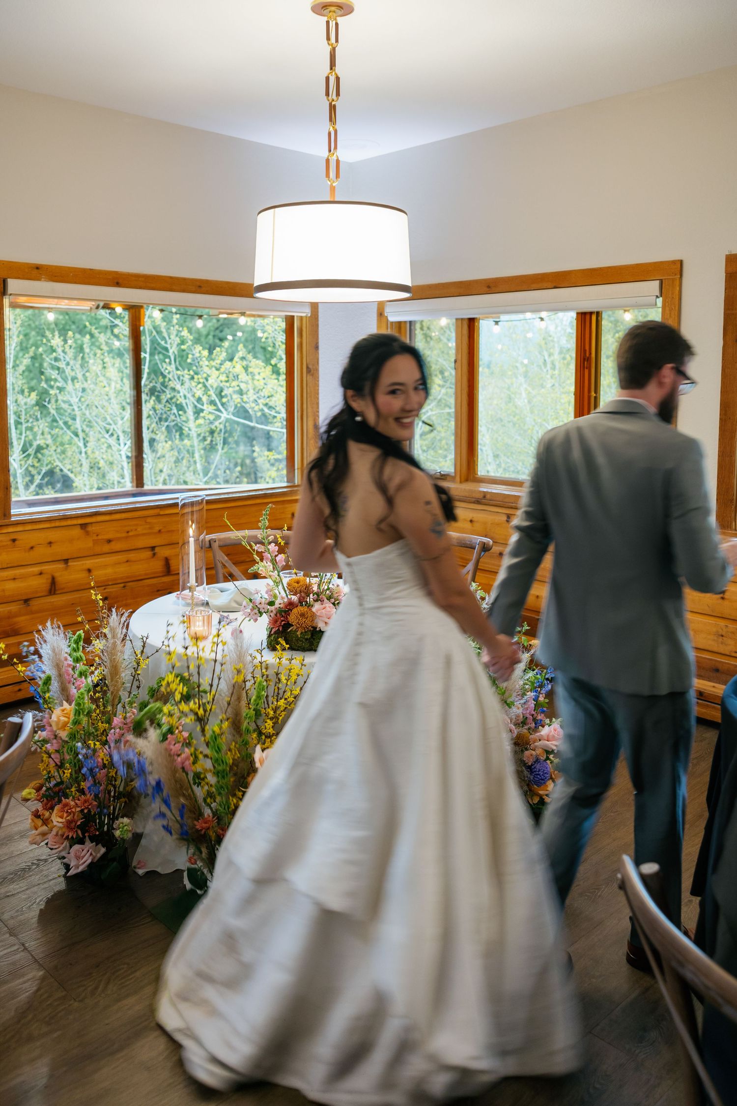 Bride in white gown smiling over shoulder while holding grooms hand in rustic wood-paneled reception venue.