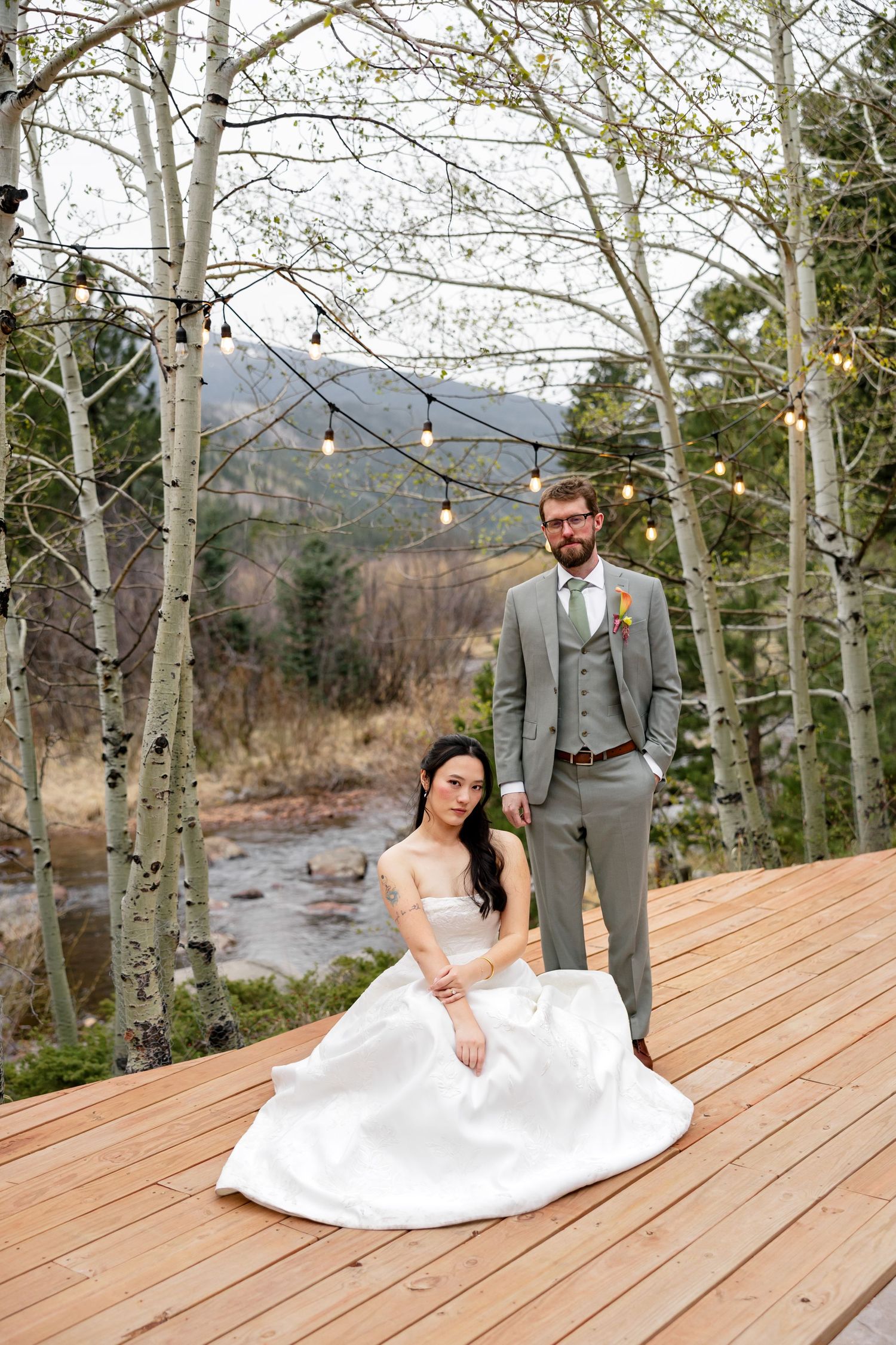 Bride in white gown sits on wooden deck as groom in gray suit stands beside her, surrounded by aspen trees and string lights.