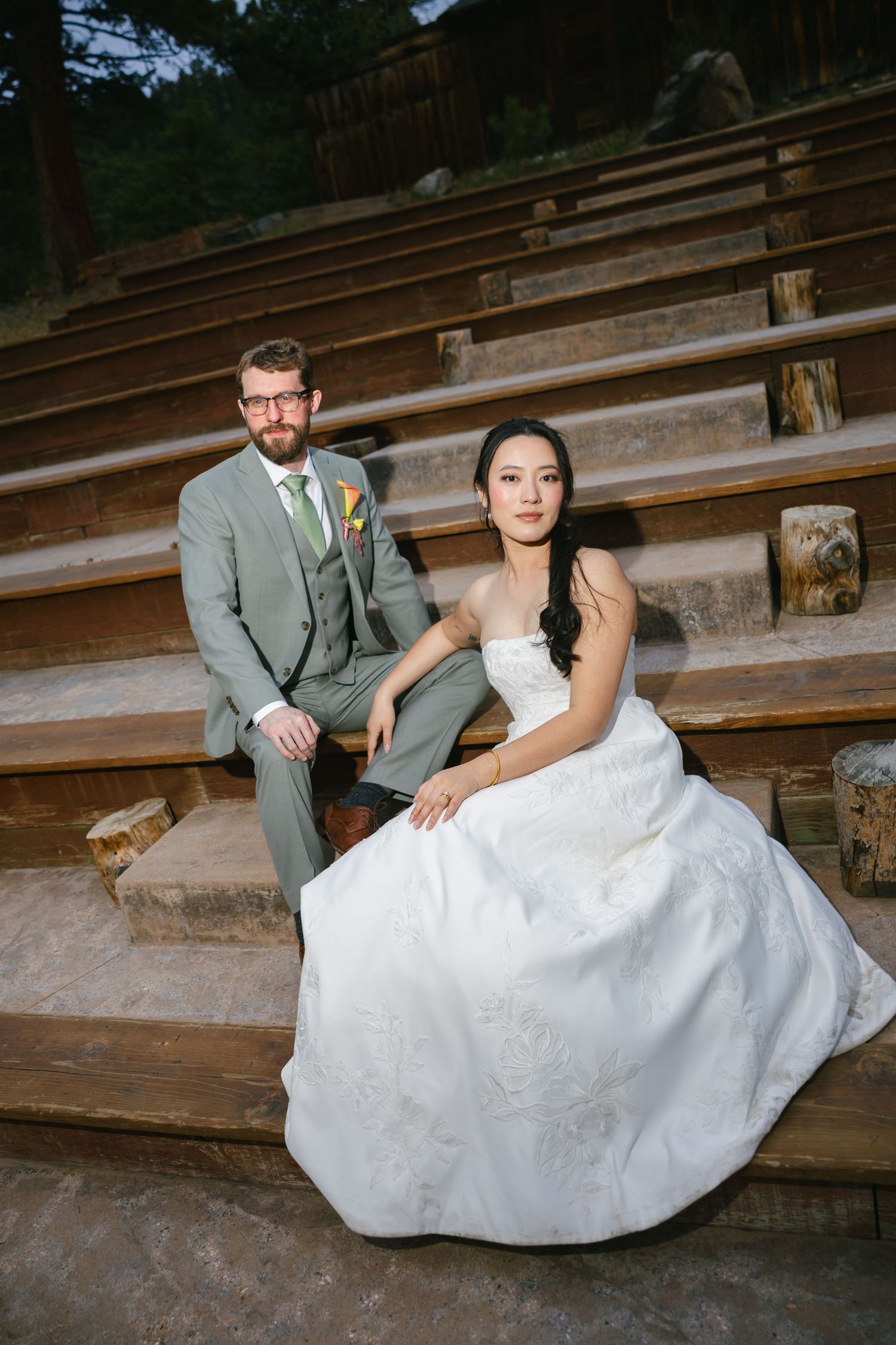 Bride in white gown and groom in gray suit pose on rustic wooden amphitheater steps at outdoor wedding.