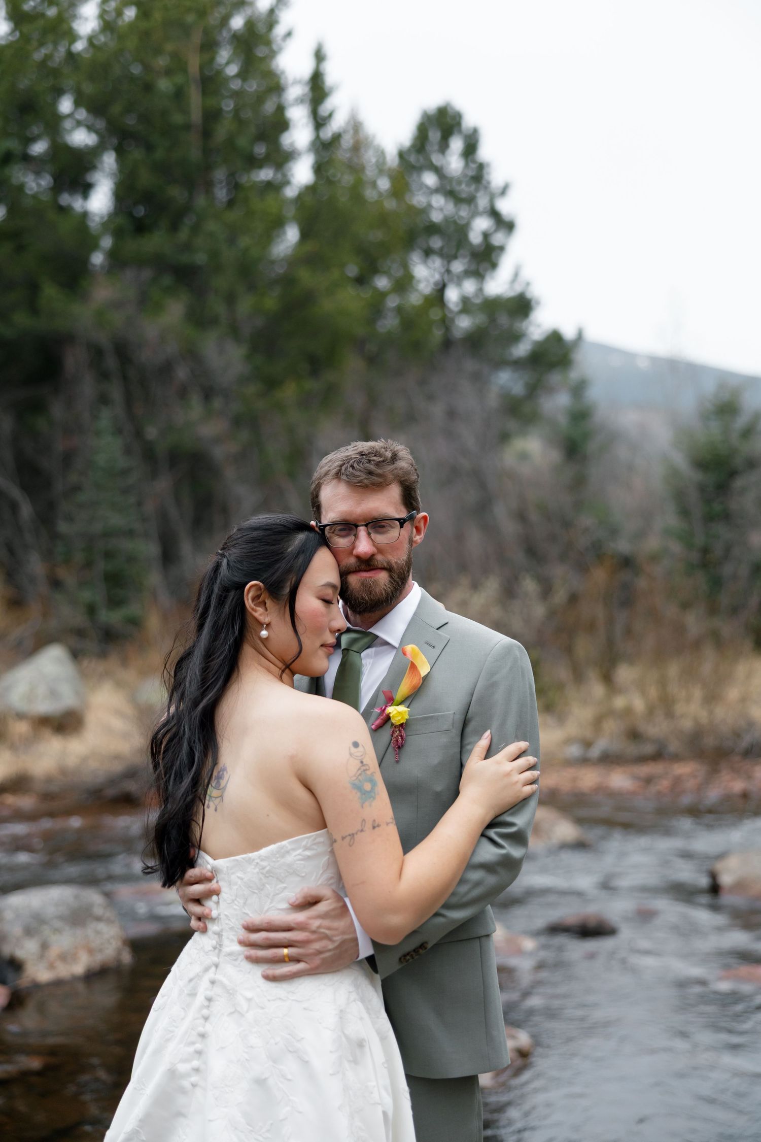 A bride and groom embrace near a mountain stream, she in a strapless lace gown, he in a gray suit with colorful boutonniere.