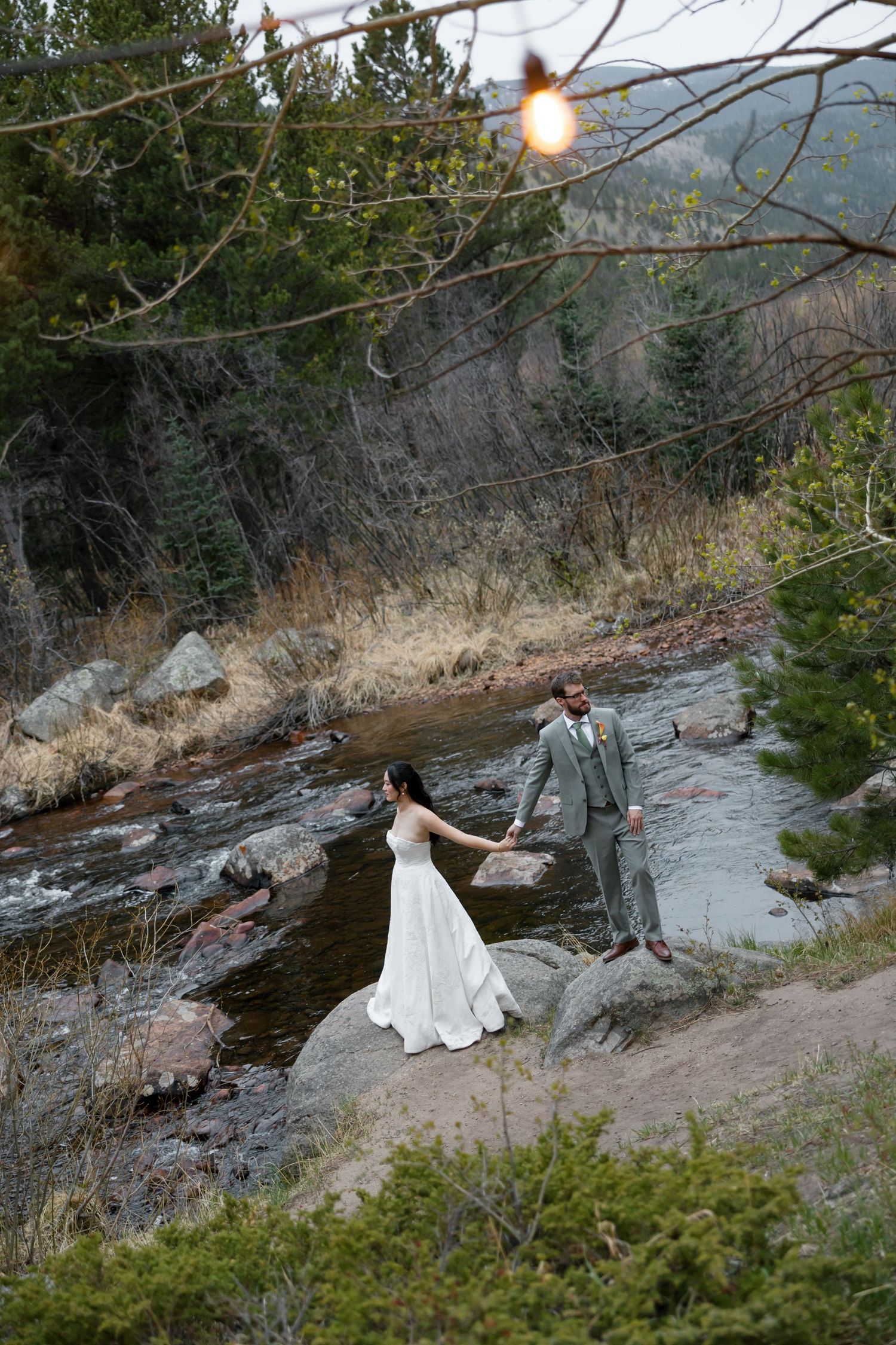 Bride and groom pose on rocky riverbank surrounded by pine trees in mountain wedding portrait.