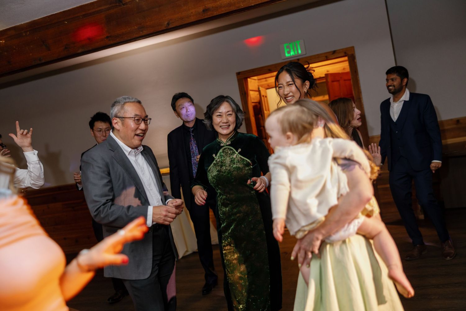 Guests dancing and laughing at a wedding reception, woman in green cheongsam with baby in foreground.