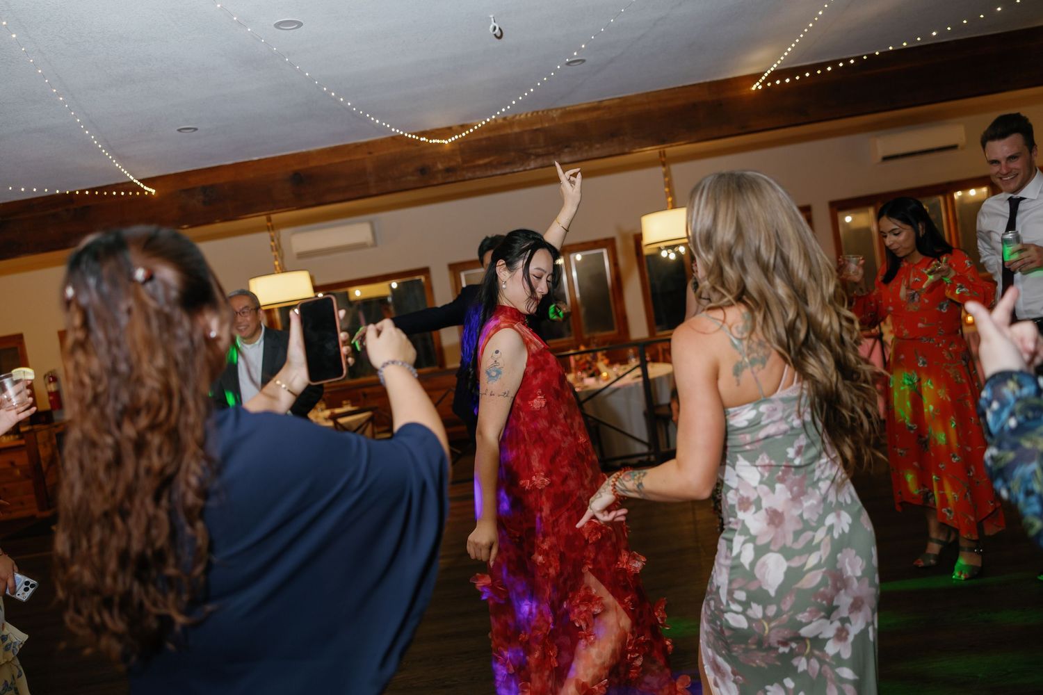People dancing in colorful dresses at an indoor wedding reception with string lights overhead.