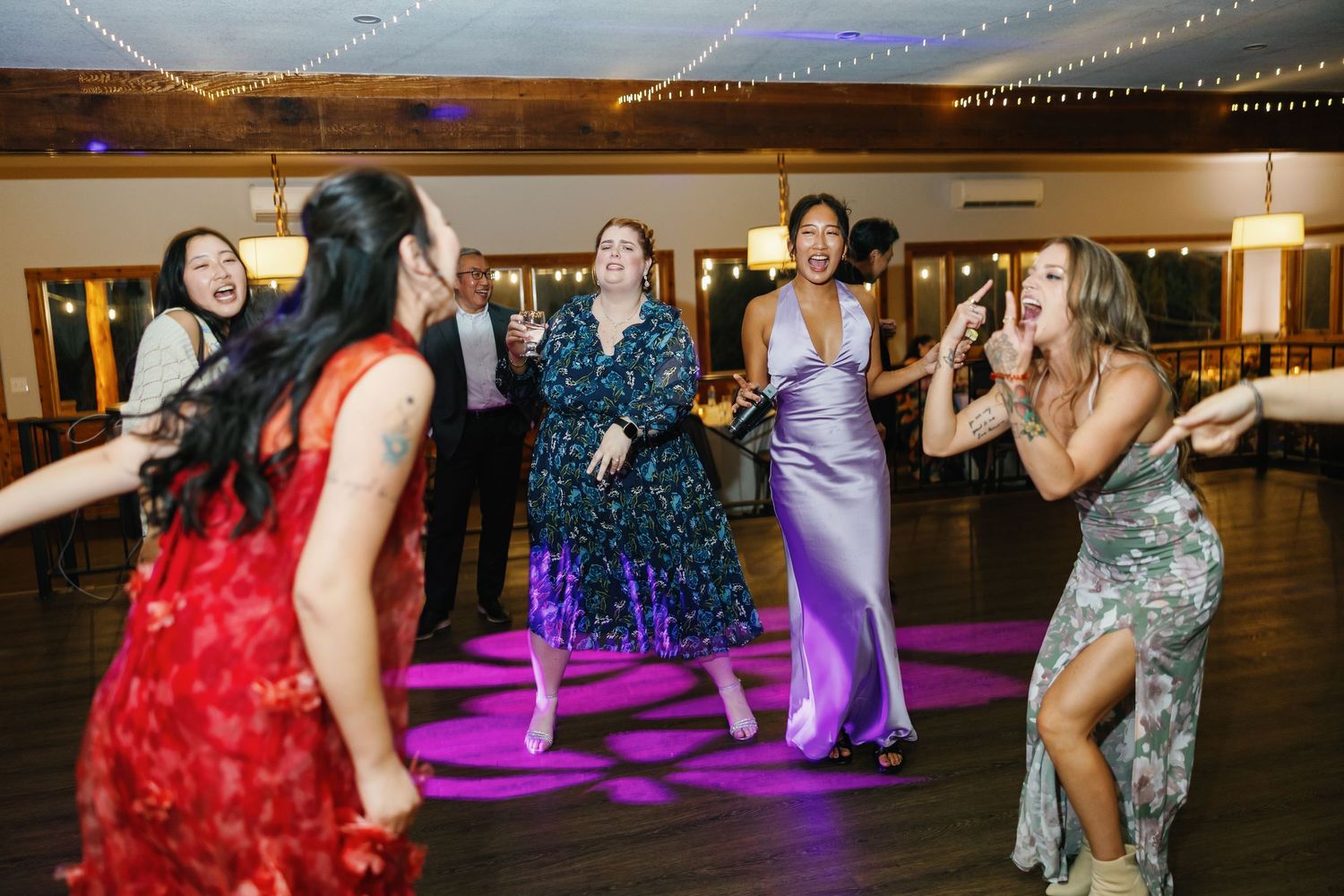 Women in elegant dresses dancing and celebrating on a purple-lit dance floor at a wedding reception.