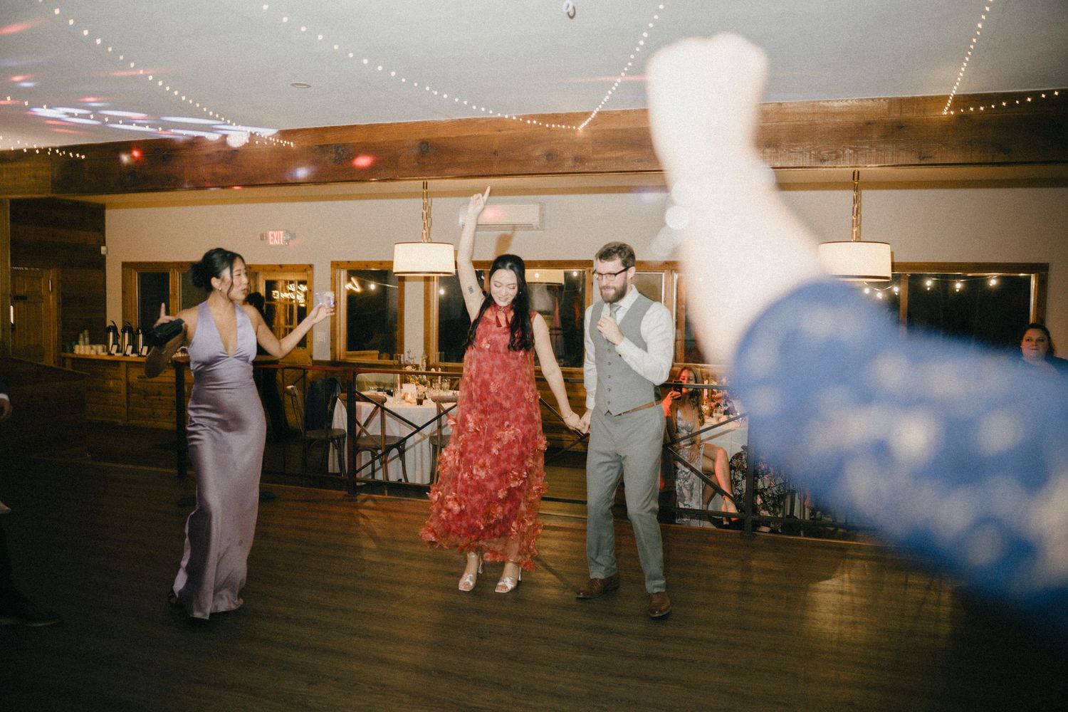 Wedding guests dance joyfully on the dance floor during a lively indoor wedding reception with warm string lights.