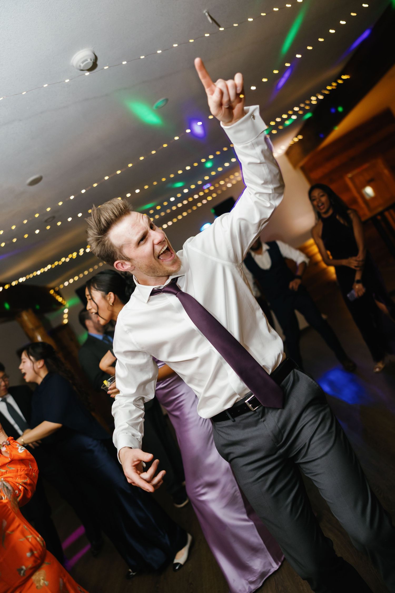 Man in white shirt and purple tie dancing energetically at a formal event with colorful lights in the background.