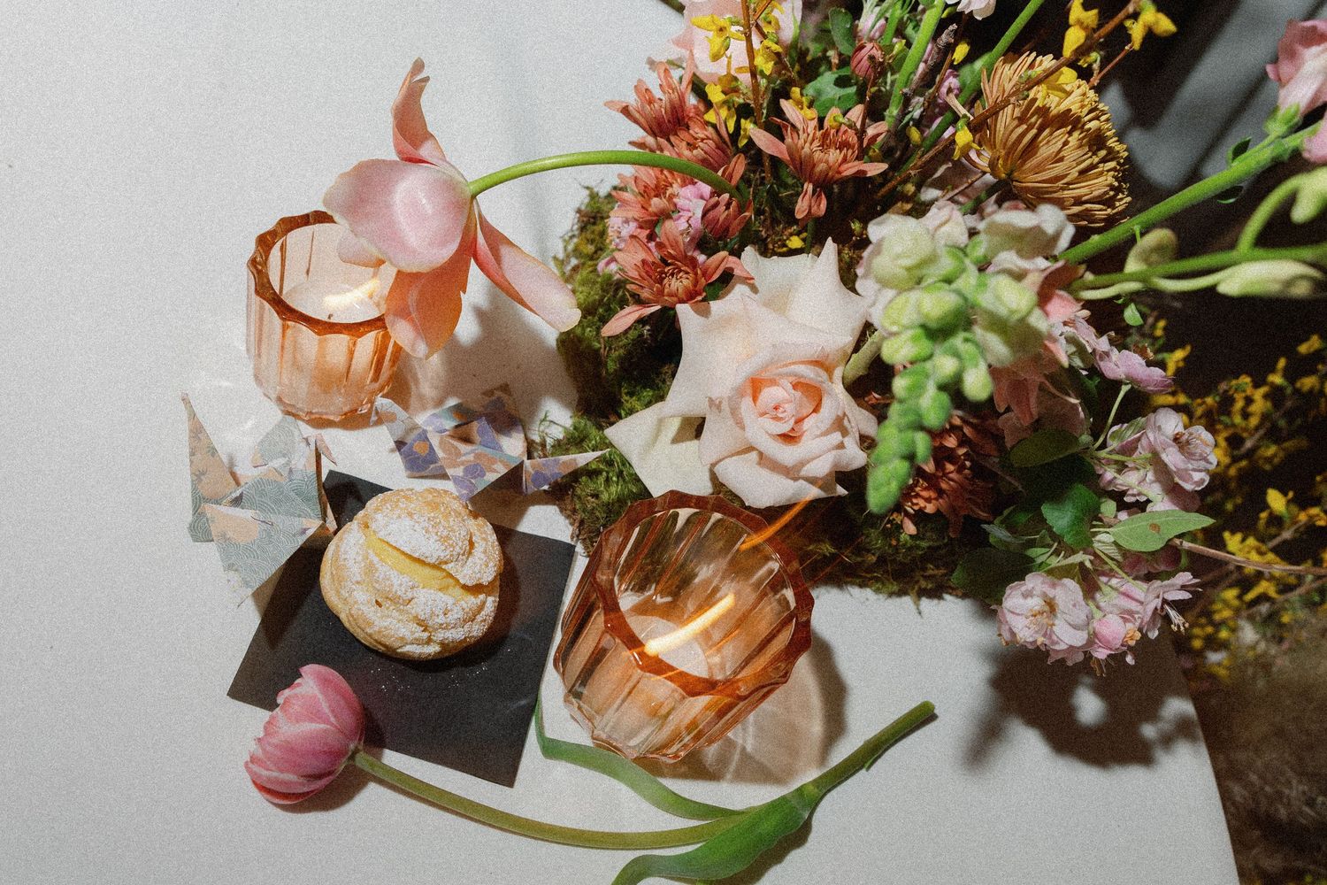 Elegant floral arrangement with roses and wildflowers beside glowing amber candles and crystals on a white surface.