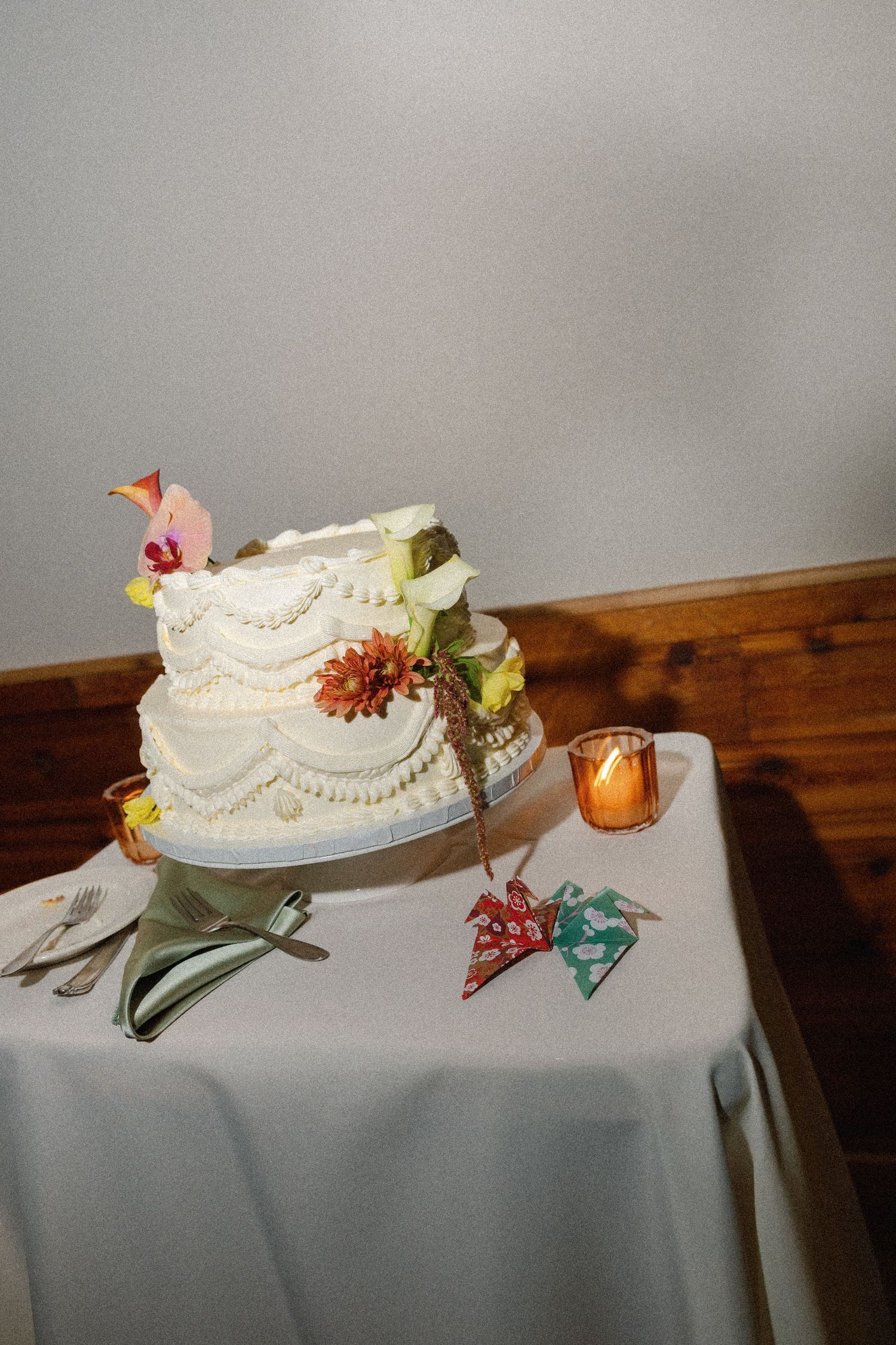 Two-tier white wedding cake with floral decorations on a draped table with candles and origami accents.