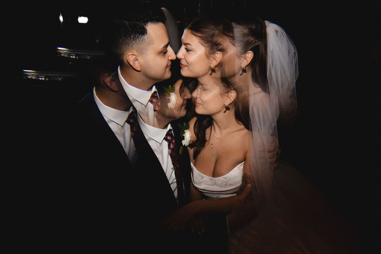 A bride and groom share a romantic moment, nearly kissing, with the bride wearing a white dress and veil in dim lighting.