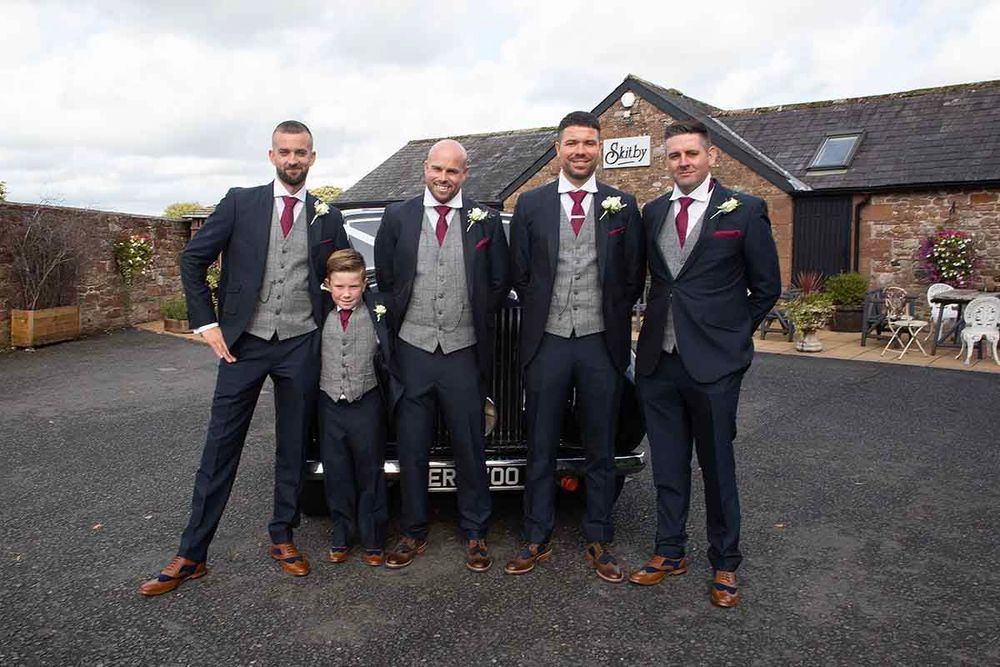 Groom and groomsmen in navy suits with grey waistcoats and red ties posing with a vintage car at  Skitby House wedding venue