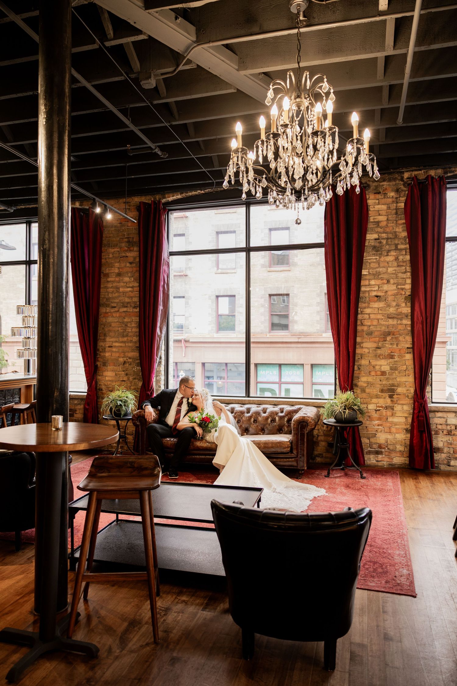 Bride and groom share an intimate moment on a leather sofa in an elegant industrial venue with chandelier and brick walls.