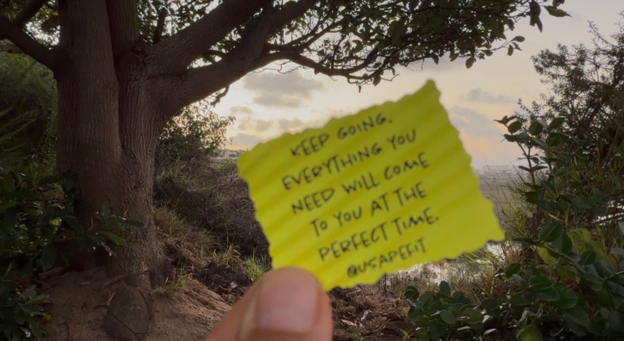 Hand holding a yellow sticky note with motivational quote among trees at sunset.