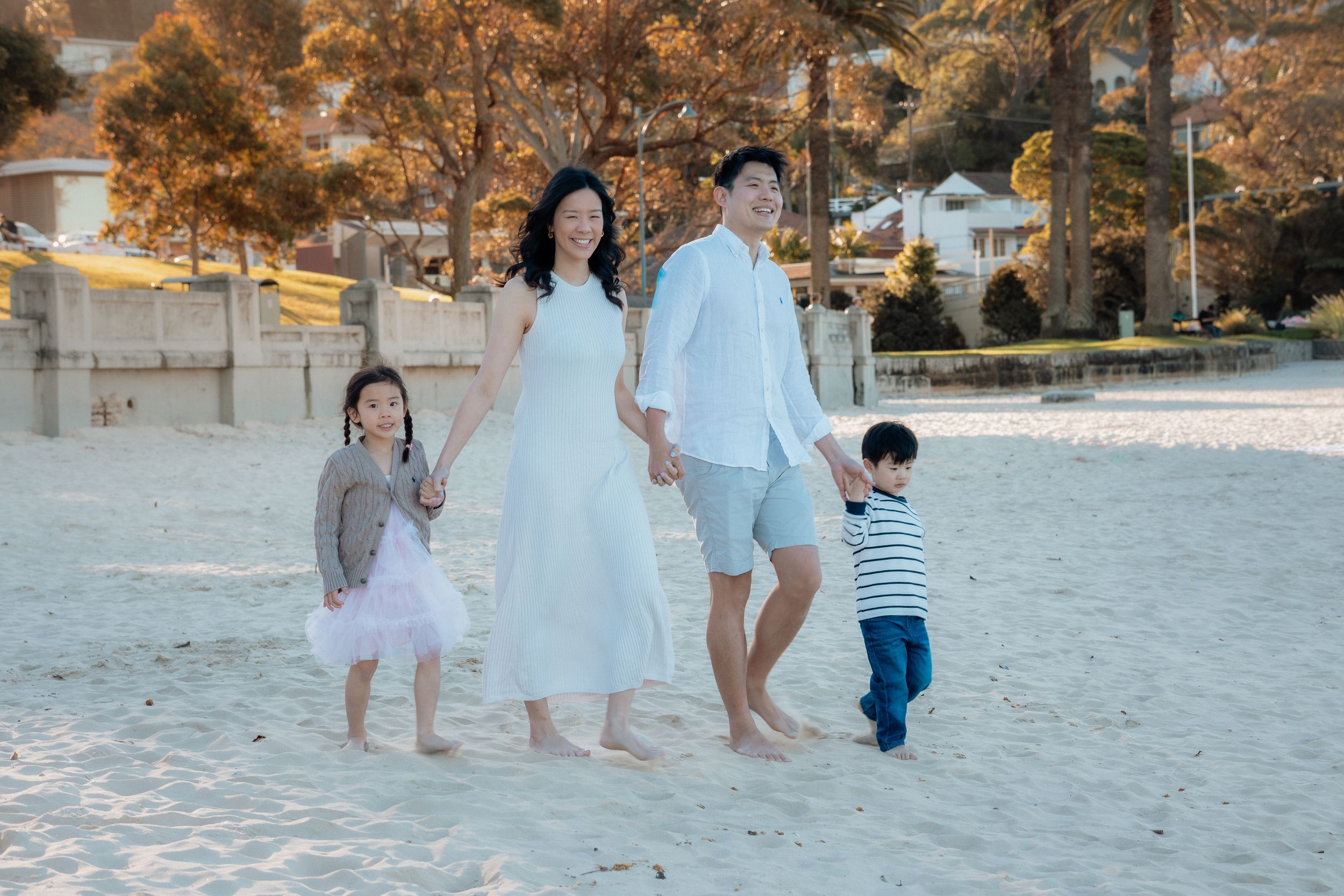 Family of four walking hand-in-hand on a sandy Balmoral beach, parents and two young children dressed in white and casual wear.
