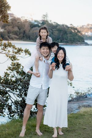 Happy Asian family of four posing outdoors near a scenic waterfront, parents carrying children on shoulders.