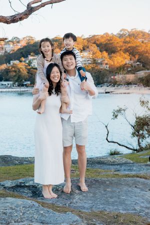 Happy Asian family of four posing barefoot on rocks by a tranquil waterway, parents carrying children on shoulders.