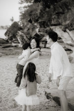 Black and white photo of a joyful Asian family laughing and embracing on a rocky beach.