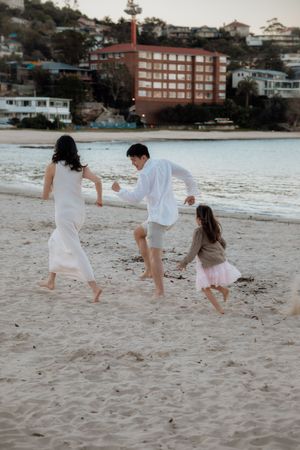 A family of three runs playfully along a sandy beach at dusk with buildings in the background.