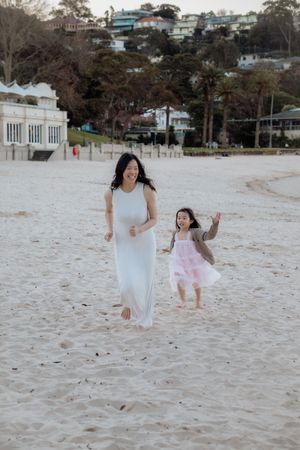 A woman in a white dress runs joyfully with a young girl in a pink tutu on a sandy beach.