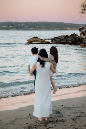 Mother with two children standing on sandy beach at sunset facing the ocean.