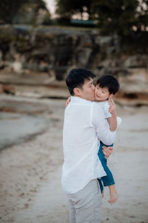 A father kisses his young son on the cheek while holding him on a sandy beach.