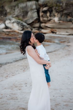 A mother in a white dress holds and kisses her young son on a sandy beach with rocky cliffs in the background.