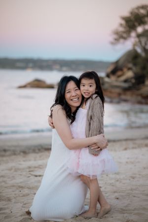 A mother and young daughter in pink dresses share a warm hug on a sandy beach at sunset.