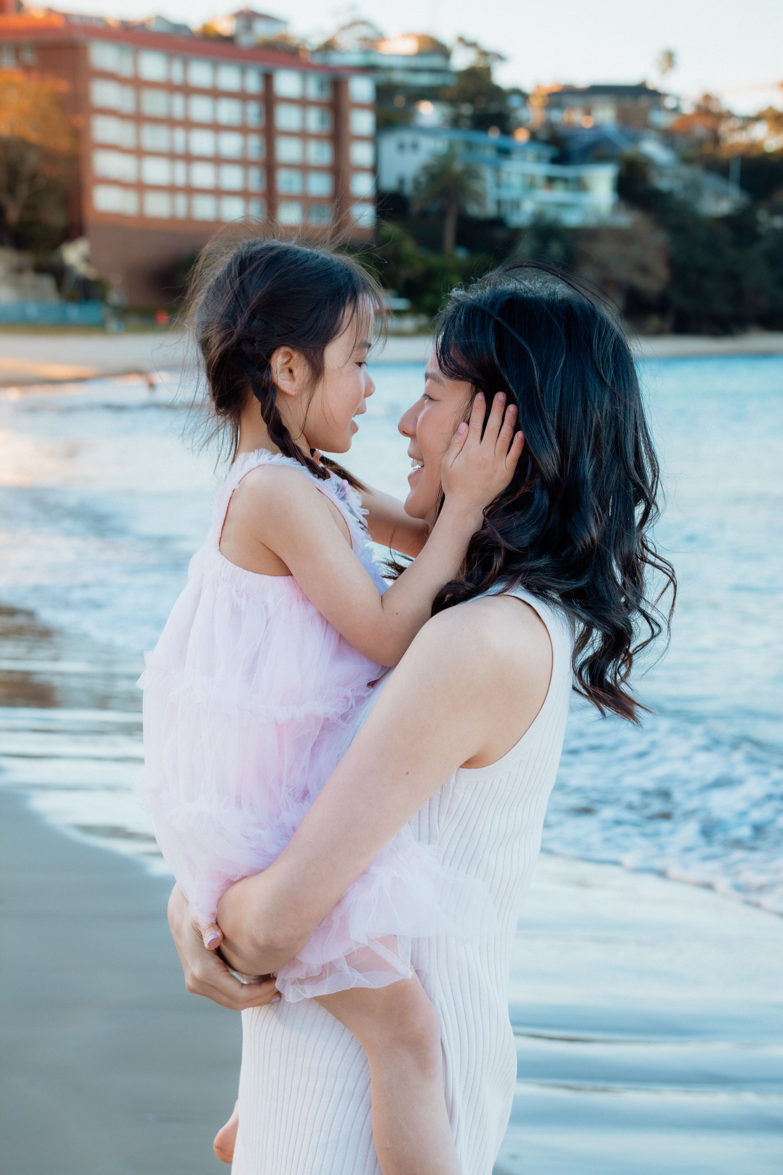 A mother holds her young daughter on a beach at sunset, sharing a tender moment with buildings in the background.