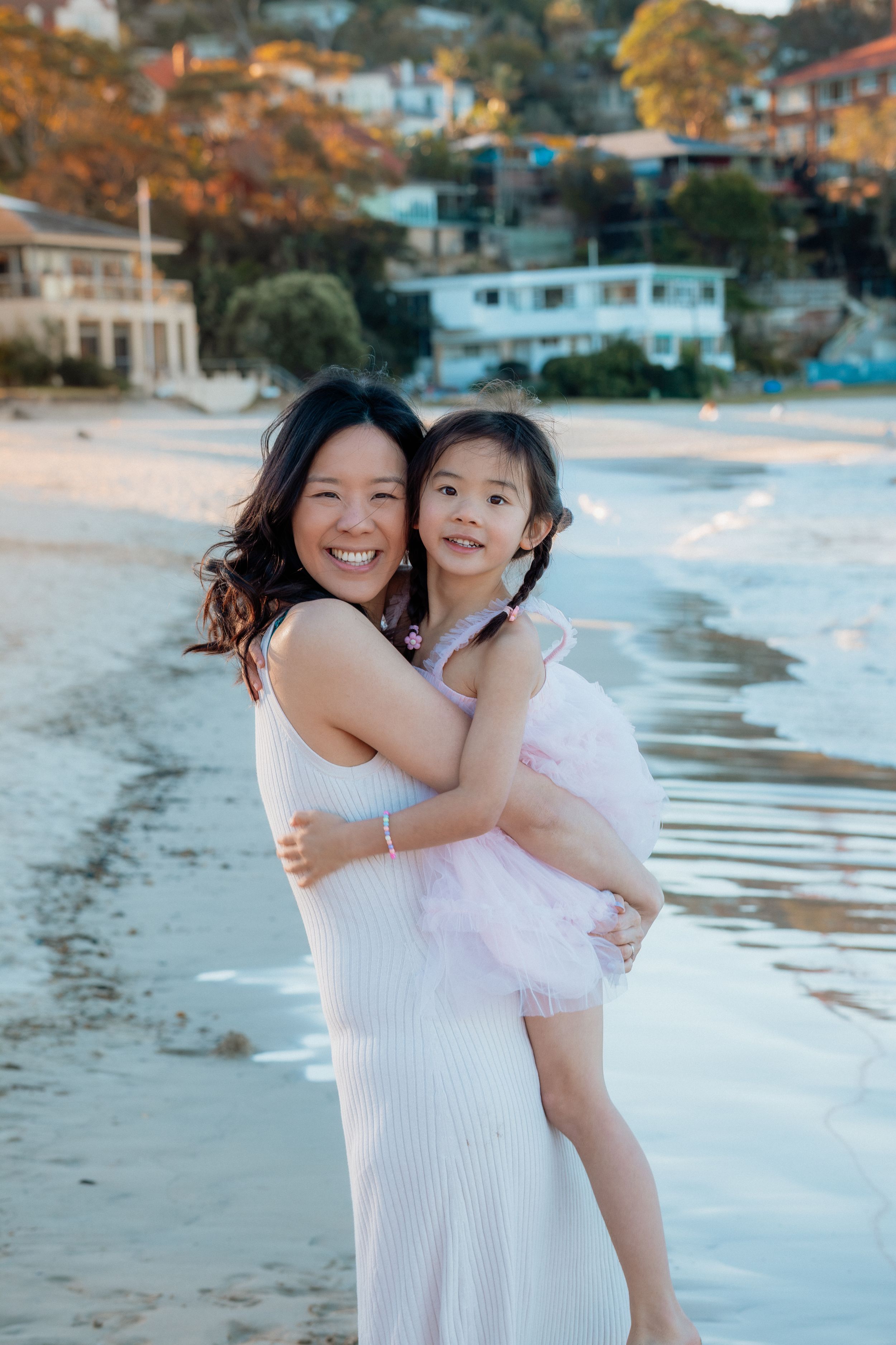 A smiling Asian woman in a white dress holds a young girl in a pink tutu on a sandy beach with houses in the background.