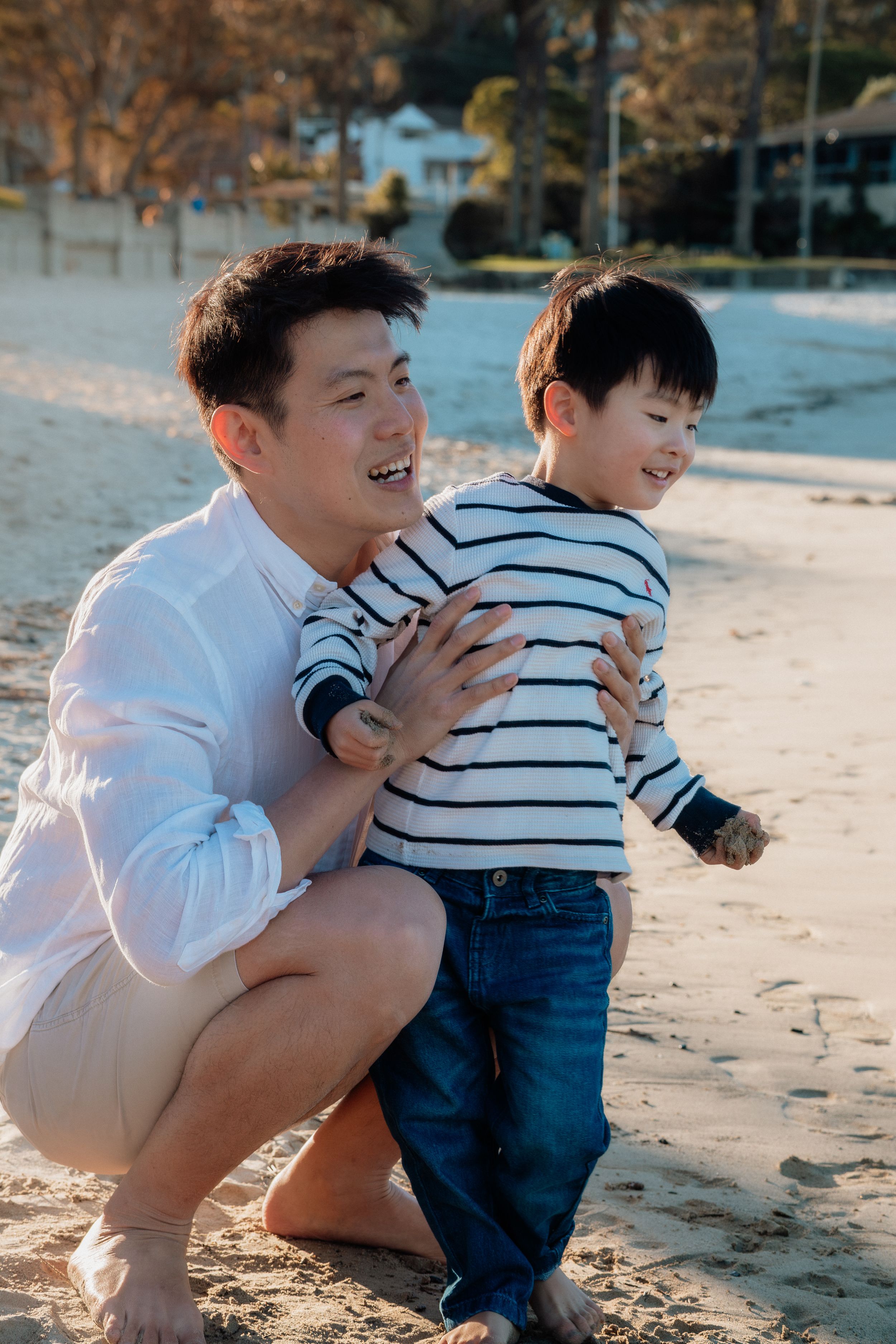 A father crouches behind his young son on a sandy beach, both smiling joyfully on a sunny day.