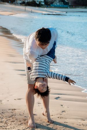 A man playfully holds a laughing child upside down on a sandy beach near the ocean waves.