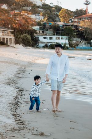 A man and young boy walk hand-in-hand along a sandy beach with coastal homes in the background.