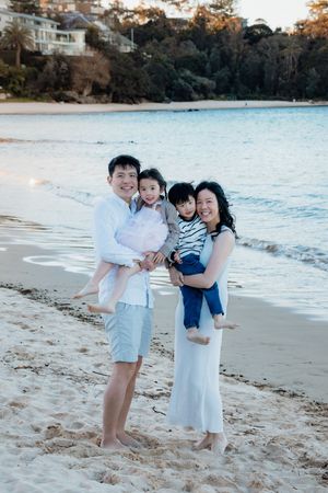 Asian family of four smiling on sandy beach at sunset, parents holding two young children, ocean and trees in background.