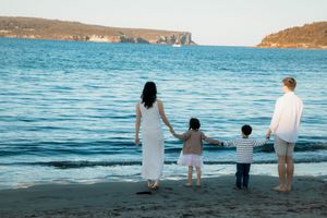 Family of four holding hands on a sandy beach, gazing at calm blue ocean waters with rocky cliffs in the distance.