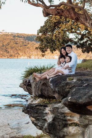 Family of four smiling on rocky waterfront under a tree at golden hour.
