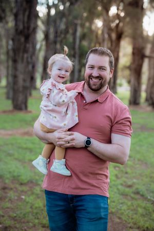 Smiling bearded man in pink polo shirt holding a laughing toddler girl in a floral dress outdoors.