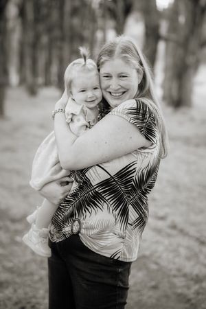 Black and white photo of a smiling woman hugging a happy toddler girl in a park setting.