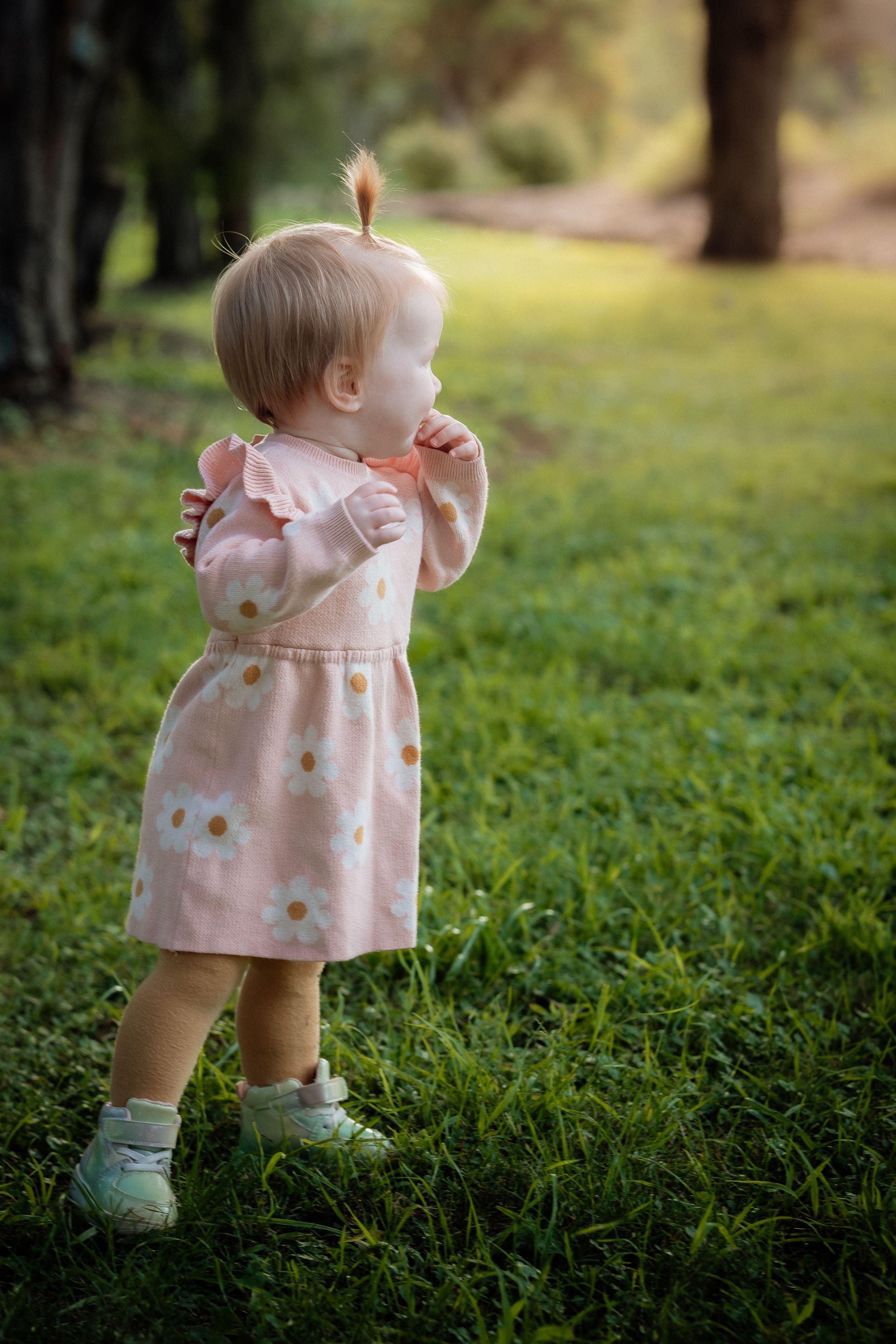 Toddler girl in pink floral dress standing on green grass in a sunlit park, looking away with hands raised.