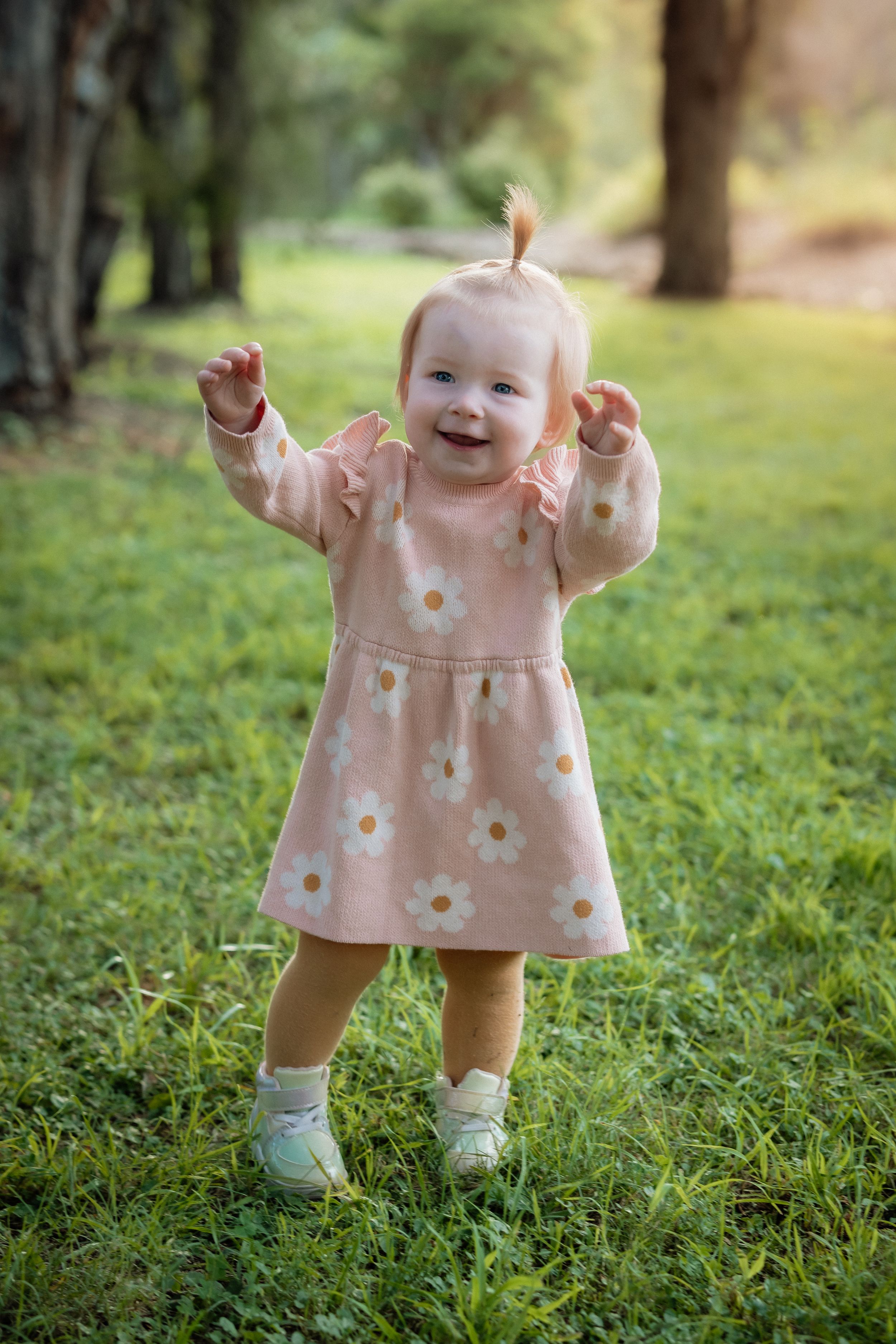 Smiling baby girl in pink daisy dress standing on green grass in a sunny park.