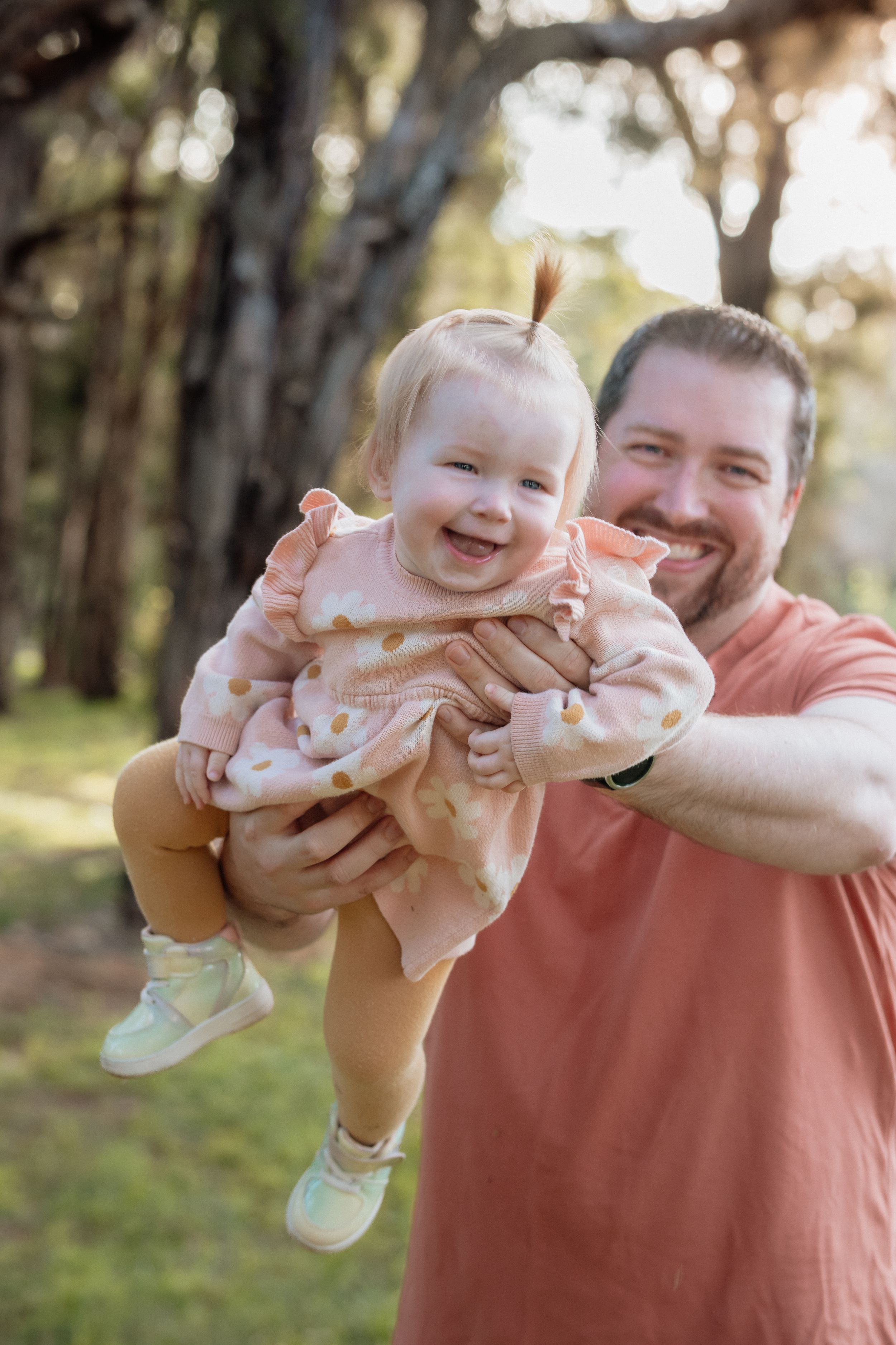 Happy father holding laughing baby girl in pink polka dot dress outdoors in sunlit park setting in Beaumont Hills