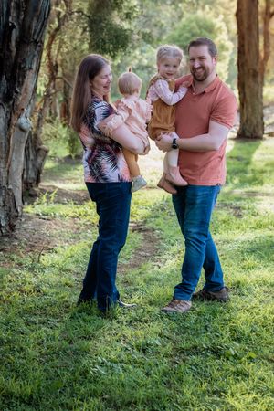 A happy family of four poses outdoors in a lush green forest, parents holding two young toddlers while smiling in Beaumont Hills