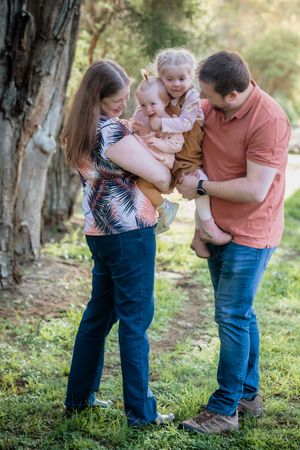A young family with two small children share a joyful moment outdoors in a green park setting.