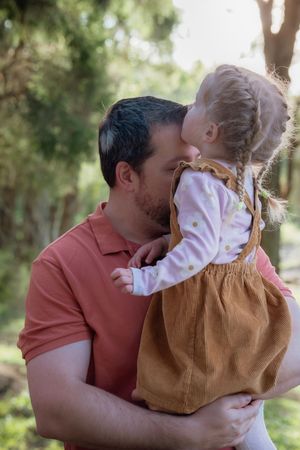 A father holding and kissing his young blonde daughter outdoors in a green park setting.