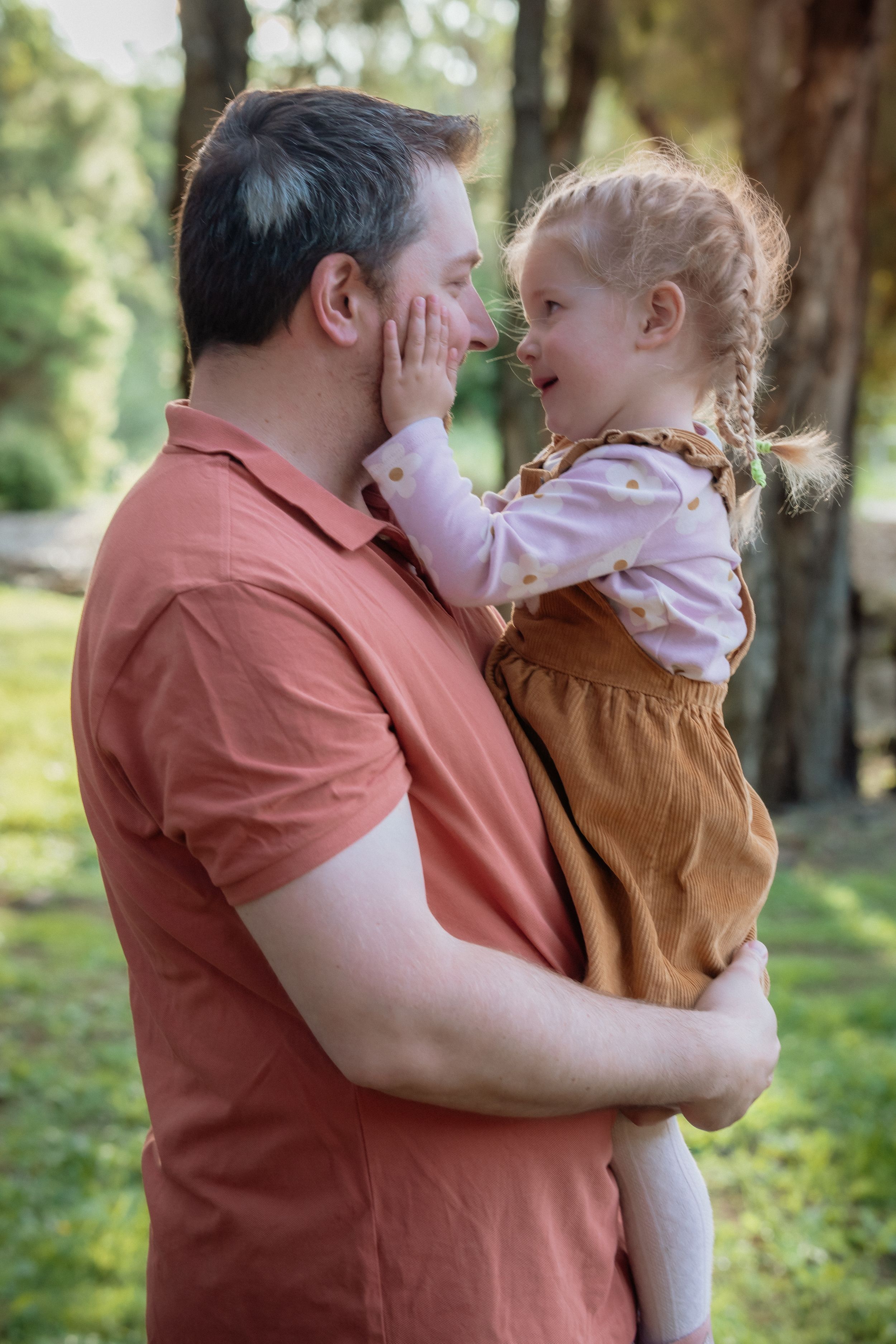 A father holds his laughing toddler daughter outdoors, as she gently touches his face in a park.