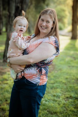 Smiling woman holding a baby in a park, both wearing casual clothes, surrounded by green trees.