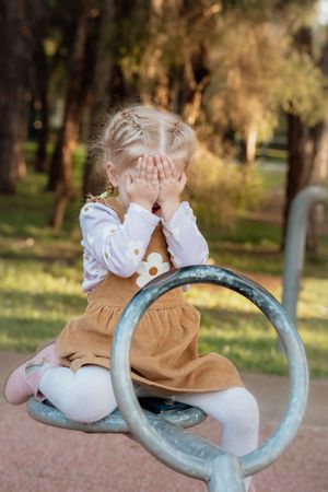Young blonde girl in brown dress covering face with hands while sitting on playground equipment outdoors.