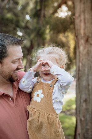 A smiling father holds his toddler daughter in a daisy dress as she makes a heart shape with her hands outdoors.