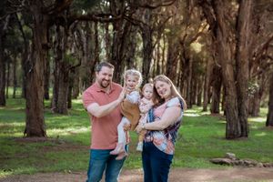 Happy family of four posing outdoors in a lush green forest, parents holding two young toddlers on a dirt path.