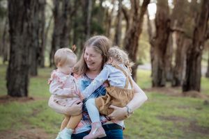 A smiling mother holds two young toddlers in her arms outdoors among tall trees in a park.