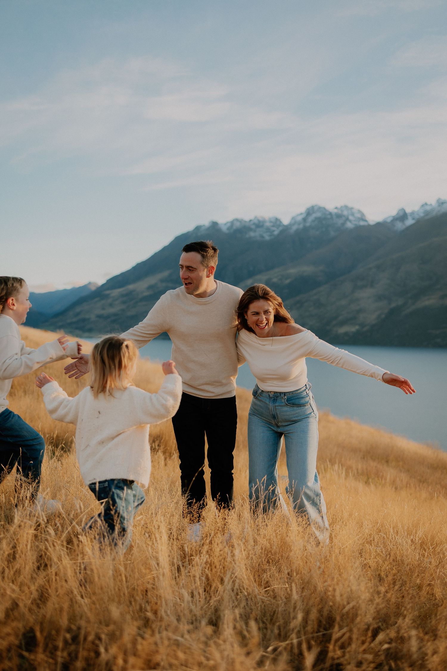 Family playing in golden grass field with mountain lake backdrop on sunny day.